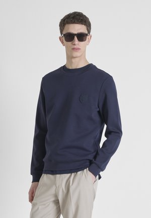 Sudadera azul marino con cuello redondo, mangas largas y un emblema de oso texturizado en el pecho. Combinada con pantalones beige.