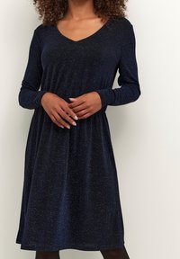 Dlouhé rukávy, šaty s V-výstřihem vyrobené z pružné, tmavě námořnické látky s metalickým leskem. Mají volný střih a na bocích jsou nařasené v pase.