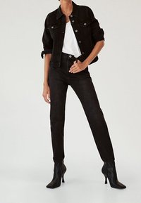 Chaqueta de mezclilla negra con botones de metal, combinada con una camiseta blanca y jeans negros de talle alto. Modelo lleva botas de tacón negro con punta afilada.