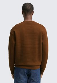 Felpa a girocollo marrone con texture, dotata di polsini e orlo a coste. Presenta un piccolo etichetta nera sulla manica sinistra. Indossata con jeans blu.