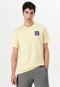 Camiseta de algodón amarilla con cuello redondo, mangas cortas y un cuadrado azul con un logo blanco "cK" en el pecho.