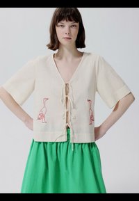 Žena s kratkom smeđom kosom nosi kremastu bluzu s vezom crvenih ptica i svijetlosmeđu zelenu suknju, stojeći s rukama na bokovima.