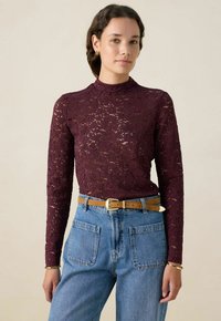 Burgunderfarbenes Spitzenoberteil mit langen Ärmeln und hohem Kragen, kombiniert mit hellblauen, hoch taillierten Jeans und einem hellbraunen Gürtel. Verfügt über Blumenmuster.