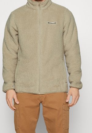 Beige Columbia Fleecejacke mit durchgehendem Reißverschluss, getragen mit brauner Cargohose, gezeigt von Hals bis Mitte Oberschenkel, Hände entspannt an den Seiten.