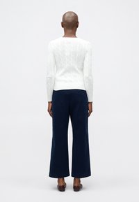 Personne debout, vue de dos, portant un pull en maille torsadée blanc, un pantalon large bleu marine et des chaussures plates marron sur un fond uni.