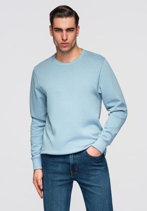 Junger Mann mit zurückgekämmtem Haar, trägt einen hellblauen Pullover und blaue Jeans, steht mit einer Hand in der Hosentasche vor neutralem Hintergrund.