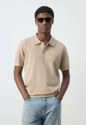 Joven con gafas de sol oscuras, polo beige de manga corta y vaqueros azul claro, de pie con las manos en los bolsillos frente a un fondo liso.