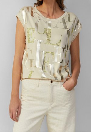 Beżowa koszula z krótkim rękawem, ozdobiona metalicznymi literami i kryształkami. Do tego jasne spodnie o fakturowanej strukturze z kieszeniami.