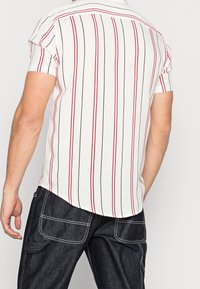 Camicia a maniche corte di colore bianco con righe verticali rosse e nere, realizzata in tessuto leggero. Presenta un colletto e un orlo curvo.