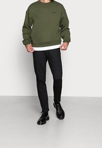 Homme portant un sweat-shirt Levi's vert olive, un sous-vêtement blanc, un jean noir et des chaussures en cuir noir, debout sur un sol gris uni.