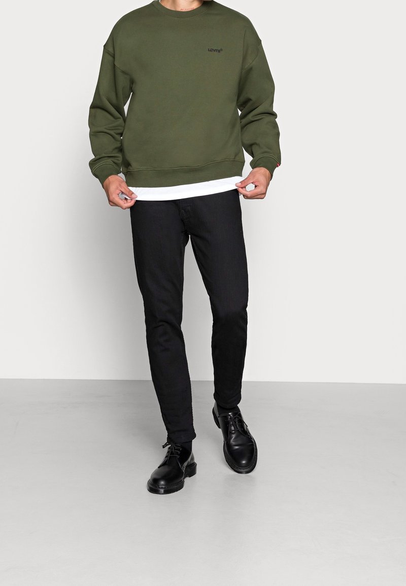 Homme portant un sweat-shirt Levi's vert olive, un sous-vêtement blanc, un jean noir et des chaussures en cuir noir, debout sur un sol gris uni.