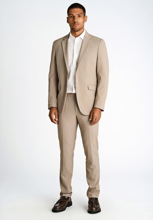 Mann steht, trägt beigen Anzug, weißes Hemd und dunkelbraune Leder-Tassel-Loafer vor einfachem weißem Hintergrund.