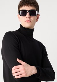 Pulover negru cu guler înalt, design ajustat, guler curbat, purtat de un model cu păr creț. Ochelari de soare mari, negri, cu rame dreptunghiulare.