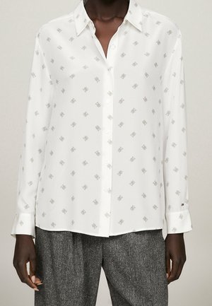 Biała koszula na długi rękaw z subtelnym wzorem logo, połączona z szarymi spodniami o fakturowanej powierzchni na osobie o ciemnej karnacji.