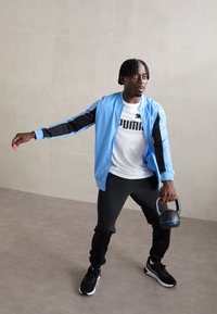 Jacheta sportivă albastră și neagră, cămașă albă cu logo negru, pantaloni negri, ținând o kettlebell neagră, într-un spațiu interior luminos.