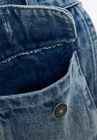 Tasca in denim con un bordo sfrangiato, dotata di un bottone argento impresso con il logo del marchio, in una sfumatura di blu medio con cuciture visibili.