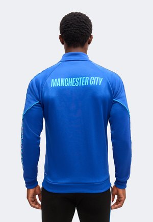 Mężczyzna w niebieskiej kurtce Manchester City z wzorzystymi paskami na rękawach, stoi tyłem do widza na jednolitym tle.