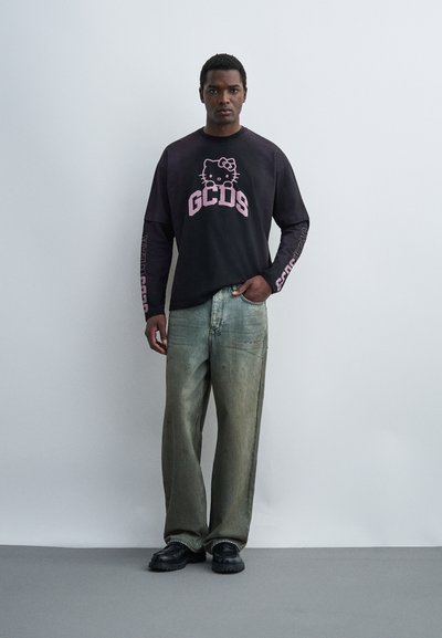 Άνδρας που στέκεται μπροστά σε απλό φωτεινό τοίχο φορώντας μαύρο πουκάμισο μακρυμάνικο με ροζ Hello Kitty και λογότυπο "GCDS", φαρδιά πράσινα παντελόνια, μαύρα παπούτσια.