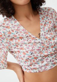 Haut enveloppant fleuri avec un design en V. Le matériau est léger, avec un fond blanc, orné de motifs de fleurs orange, rose et teal.