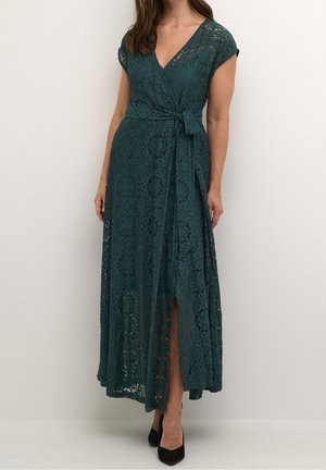 Femme portant une robe portefeuille en dentelle vert foncé avec manches courtes, encolure en V, fente latérale, et des escarpins noirs à bout pointu, debout devant un fond uni.