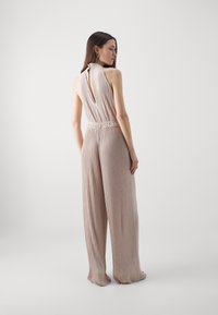 Beige sleeveless jumpsuit with vertical pleats, high neck, and exposed back detail. Loose, wide-leg fit, textured fabric.