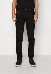 Jeans en denim noir à coupe slim, avec un petit logo brodé sur la poche avant et des détails de couture subtils. Portés avec des baskets noires.