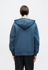 Sweat à capuche de couleur bleu doux, avec une coupe décontractée, des poignets côtelés et une texture lisse, porté avec un pantalon noir.