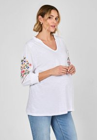 Weißes, kurzärmeliges Oberteil mit V-Ausschnitt, floralem Stickerei an den Ärmeln, Rüschensäumen, kombiniert mit hellblauen Jeans. Glattes, leichtes Material.