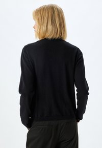 Maglione nero a maglia con maniche lunghe, scollatura rotonda e orlo a costine. Tessuto liscio; design minimalista, indossato sopra pantaloni scuri.