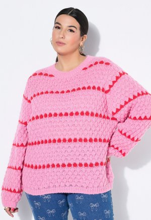 Kvinde med mørkt hår iført en lyserød strikket sweater med røde striber og blå jeans dekoreret med hvide broderede sløjfer, stående mod en hvid væg.