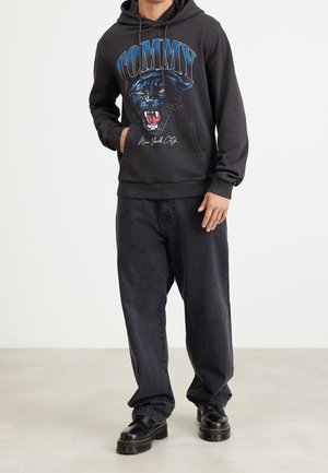 Felpa nera con cappuccio raffigurante un giaguaro ruggente, con la scritta "TOMMY" e "New York City" stampata sotto, abbinata a jeans scuri larghi e stivali neri.