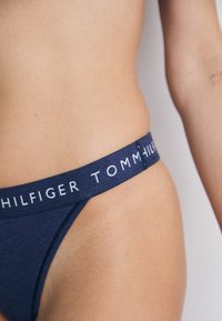 Artimo plano vaizdas, kuriame matyti asmens apatinė torso dalis, vilkinti tamsiai mėlynus Tommy Hilfiger apatinius drabužius su matoma prekės ženklo juosta.