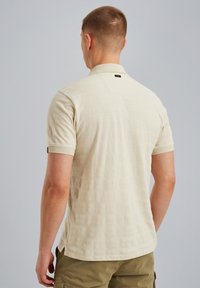 Beige kortærmet polo shirt med et struktureret mønster, en flad krave og et logo mærke på bagsiden, parret med lyse olivengrønne shorts.