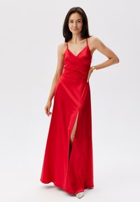 Červené saténové šaty s překládaným designem, které mají tenké ramínka a rozparek až k stehnům. Látka má hladký, lesklý povrch.
