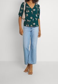 Grünblumige Bluse mit V-Ausschnitt und Puffärmeln, kombiniert mit hellblauen weit geschnittenen Jeans und beigen Riemchensandalen. Trägt eine leichte Handtasche.