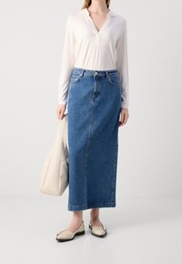 Falda larga de mezclilla en azul, de cintura alta con bolsillos frontales. Combinada con una blusa blanca de manga larga y unas bailarinas crema. Bolsa de mano beige presente.