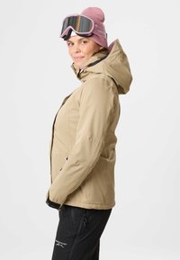 Kvinde iført beige vinterjakke, lyserød beanie og lyserøde skibriller, stående i sideprofil mod en ensfarvet baggrund.