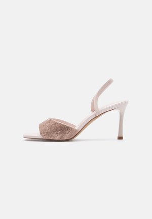 Beige sandal med høj hæl, tynd ankelrem og tæppe af glitrende rhinsten på tåremmen, vist fra siden på hvid baggrund.