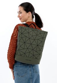 Mochila verde con un patrón geométrico hecha de material texturizado, que cuenta con un asa superior y correas de hombro ajustables. Etiqueta de marca visible.