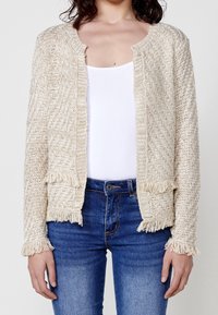 Beżowy kardigan dziany z fakturowanym wzorem, otwarty z przodu, z frędzlami przy krawędziach i mankietach. Nosi się go na białą bluzkę i niebieskie dżinsy.