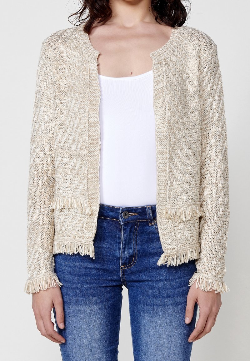 Beżowy kardigan dziany z fakturowanym wzorem, otwarty z przodu, z frędzlami przy krawędziach i mankietach. Nosi się go na białą bluzkę i niebieskie dżinsy.