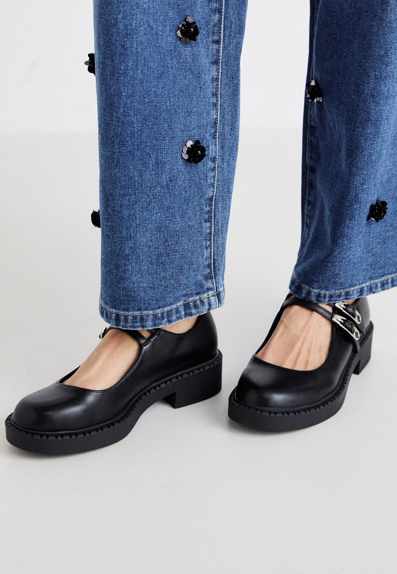 Black leather Mary Jane shoes with a square toe, chunky sole, and adjustable silver buckles, paired with blue denim pants featuring floral accents.