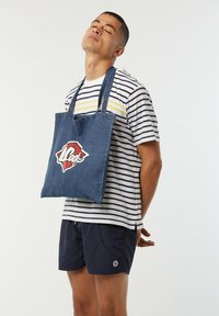 Denim-Umhängetasche aus blauem Stoff mit einem auffälligen roten und weißen Logo. Getragen über einem gestreiften Hemd und einer marineblauen Shorts.