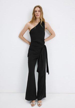 Kobieta z długimi blond włosami, ubrana w czarny top na jedno ramię z wiązanym pasem i dopasowane szerokie spodnie, stojąca na białym tle.