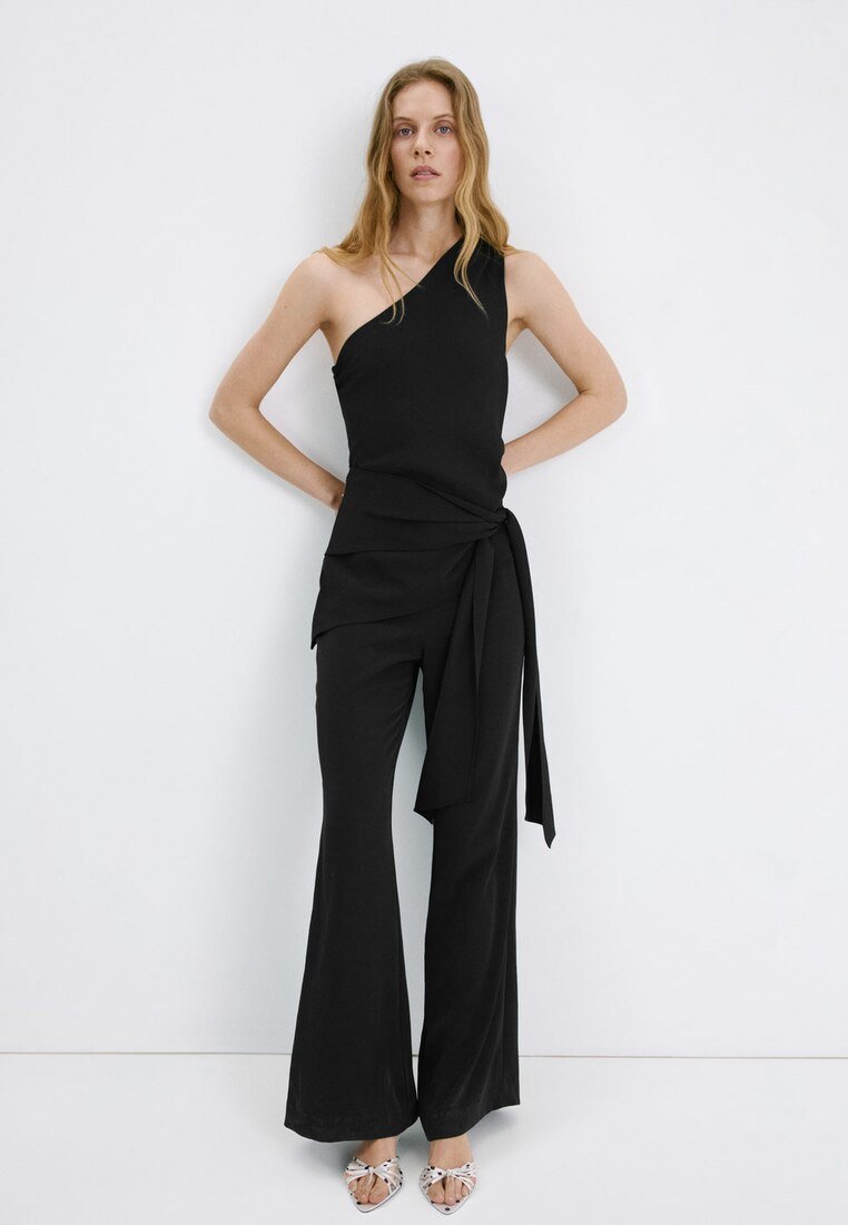 Femme aux longs cheveux blonds portant un haut noir à une épaule avec taille nouée et pantalon large assorti, debout sur un fond blanc.
