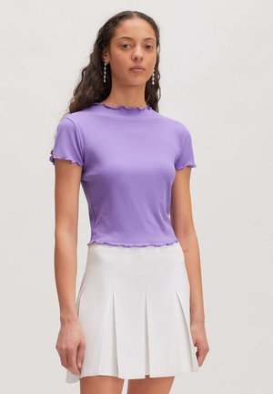 Young woman with long curly hair wearing a purple short-sleeve ribbed top and a white pleated skirt, standing against a plain background.