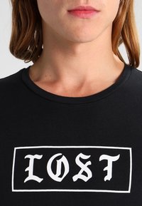 Black t-shirt featuring a white rectangular graphic with the word "LOST" in stylized font. Round neckline and soft cotton material.