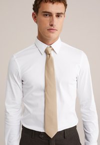 Balta ilgomis rankovėmis marškiniai su smailiu apykakle, derinami su tekstūruota šviesiai smėline kaklaraiščiu ir tamsiai pilkomis kelnėmis.