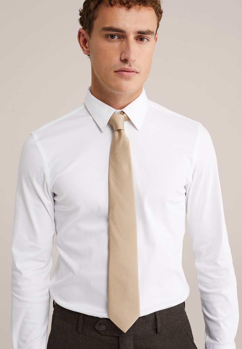 Balta ilgomis rankovėmis marškiniai su smailiu apykakle, derinami su tekstūruota šviesiai smėline kaklaraiščiu ir tamsiai pilkomis kelnėmis.