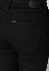 Jeans de mezclilla negros con un diseño elegante, que cuentan con un bolsillo trasero curvado y una sutil etiqueta de logo. La tela parece suave y ajustada.